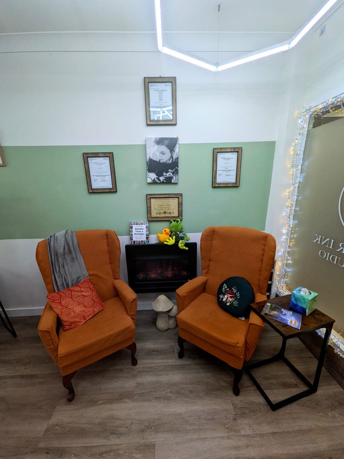 Cosy waiting corner — two orange wing-back armchairs beside an electric fire, soft neon lighting and fairy lights