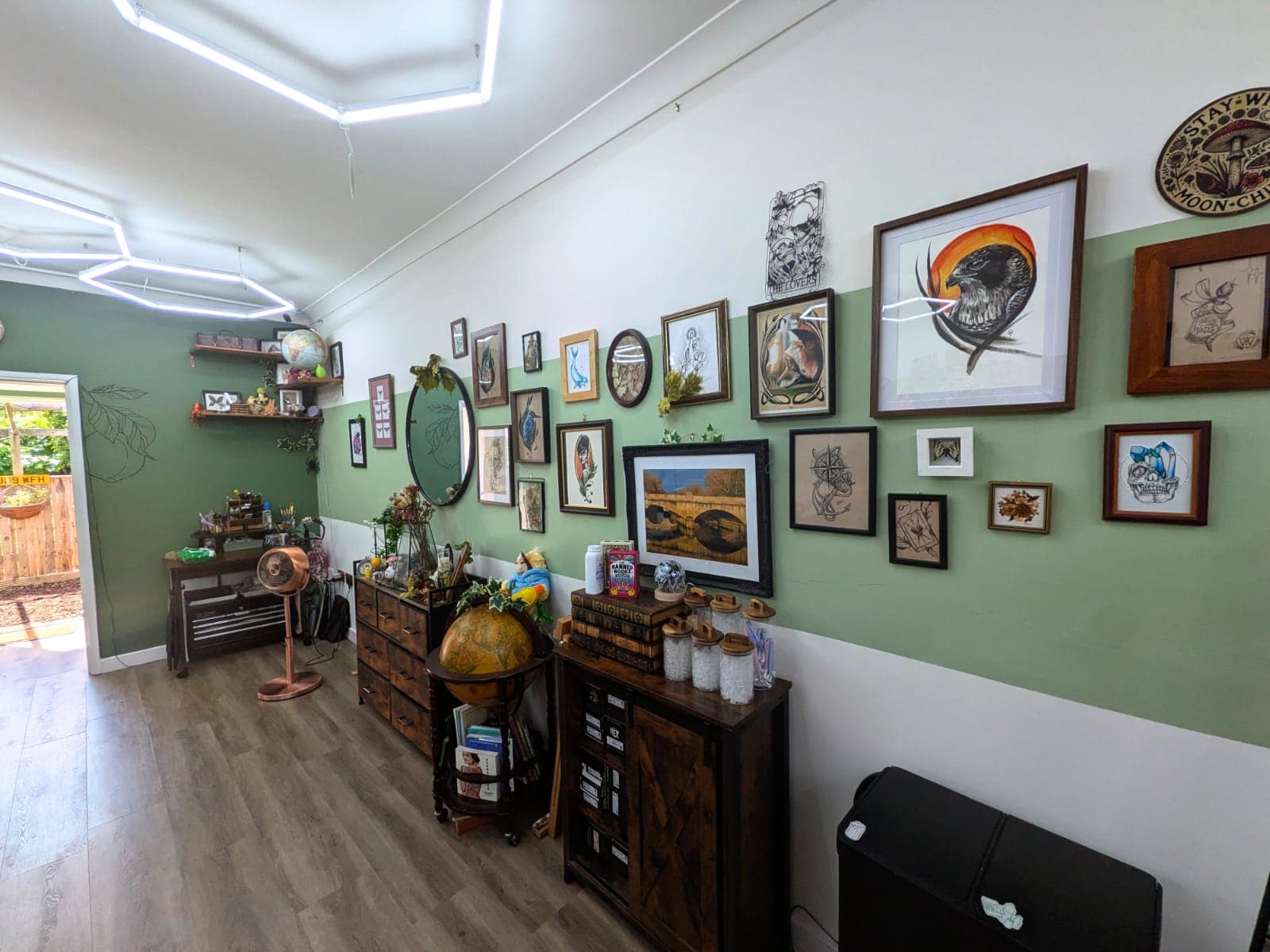 Sage-green gallery wall of framed original artwork above vintage wooden furniture and a globe