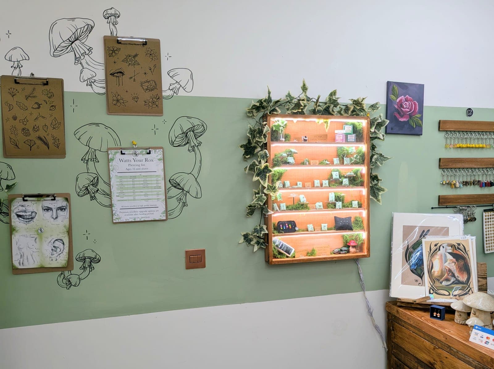 Flash designs on clipboards among hand-drawn botanical and mushroom wall murals, beside a lit shelf of plants and crystals